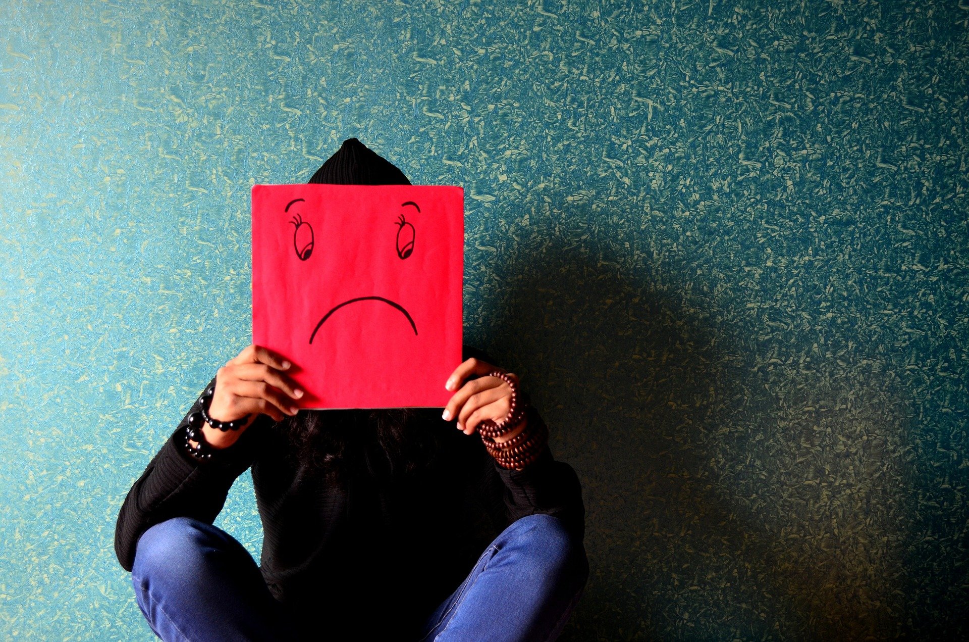 Weibliche Person sitzt vor einer Wand und hält sich ein rotes Papier vor das Gesicht, auf dem ein trauriges Gesicht gezeichnet ist.